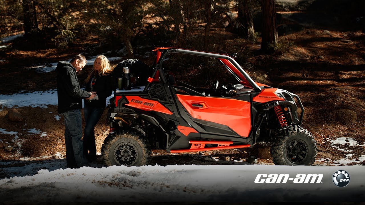 Couple standing behind their red Can-Am Maverick. Parked out in the forest with some snow on the ground.