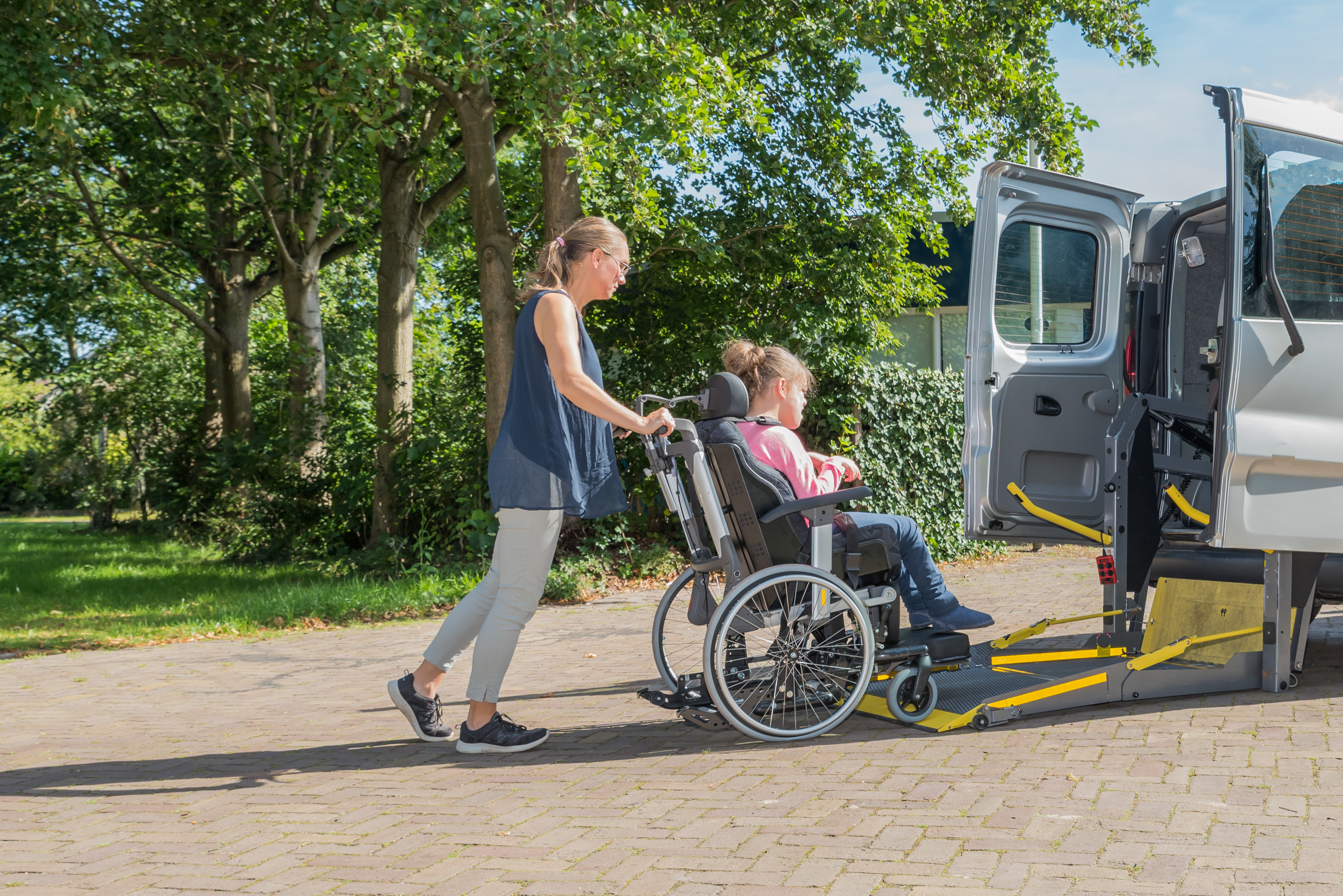 A woman pushes a younger girl in a wheelchair onto a loading platform that leads into the back of a van.
