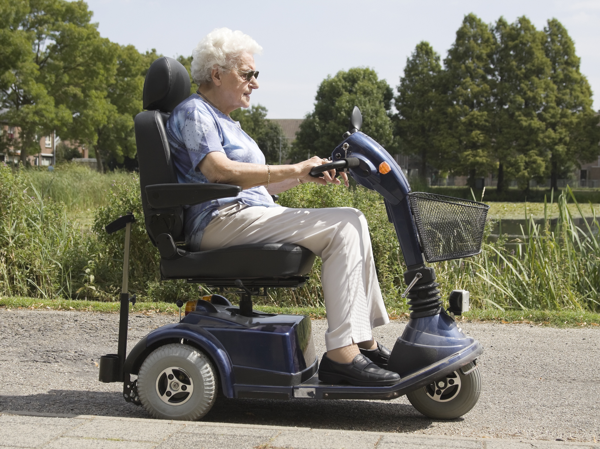 An elderly woman moving through a park on a mobility scooter.